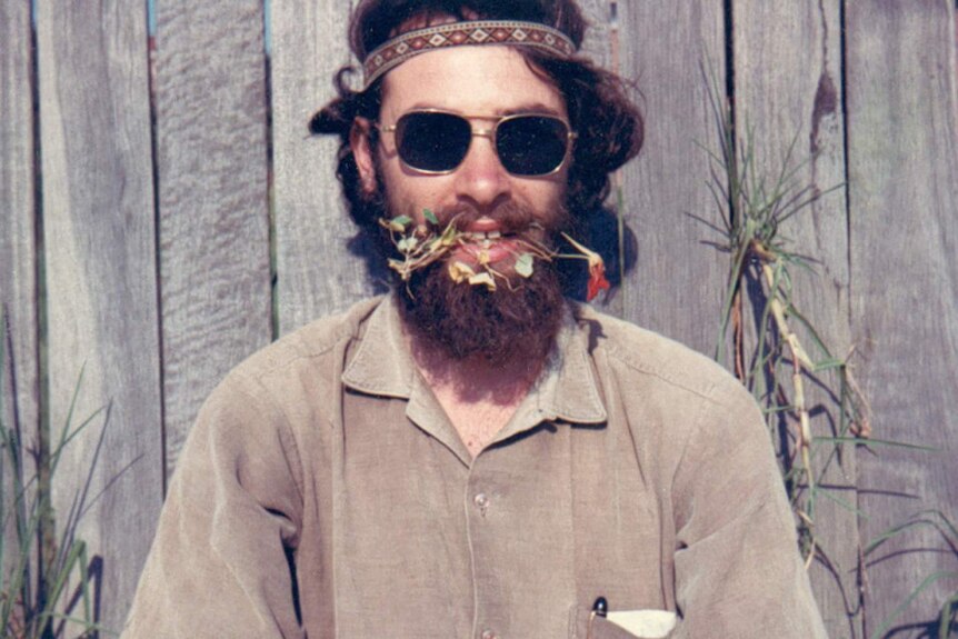 A man wearing sunglasses and a woven headband smiles with flowers hanging out of his mouth.