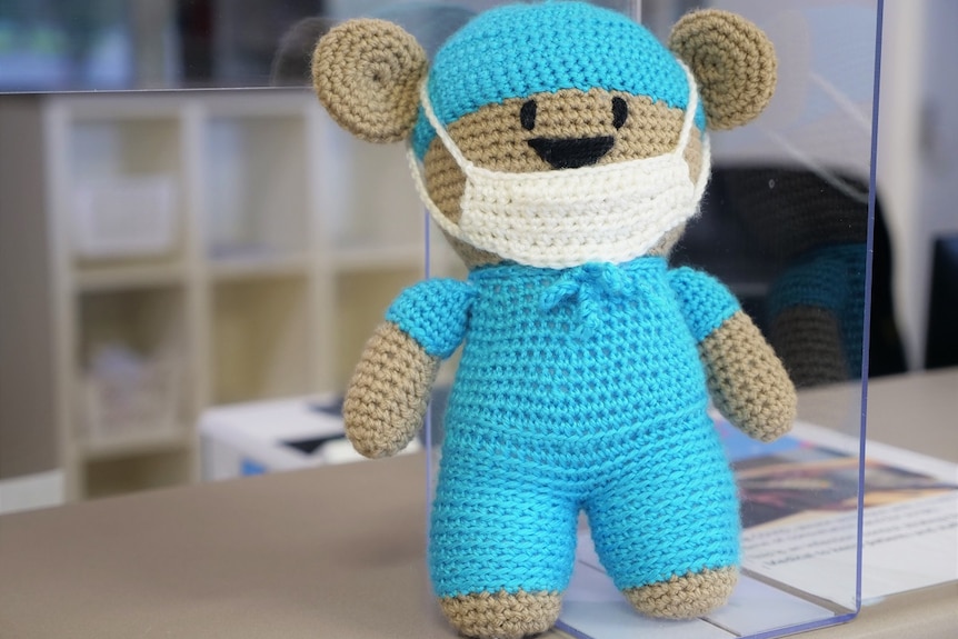 A crocheted light brown teddy bear, blue cap, blue scrubs, medical mask sitting on a reception desk.