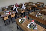 Schoolgirls sit inside a classroom with bouquets of flowers on empty desks