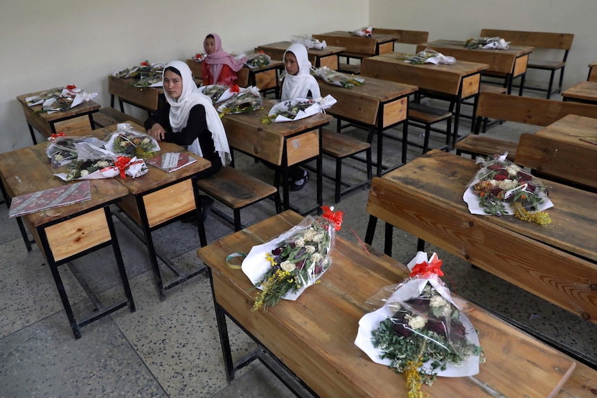 Schoolgirls sit inside a classroom with bouquets of flowers on empty desks