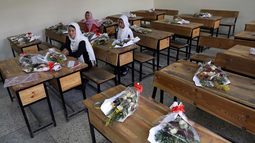 Schoolgirls sit inside a classroom with bouquets of flowers on empty desks