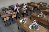 Schoolgirls sit inside a classroom with bouquets of flowers on empty desks