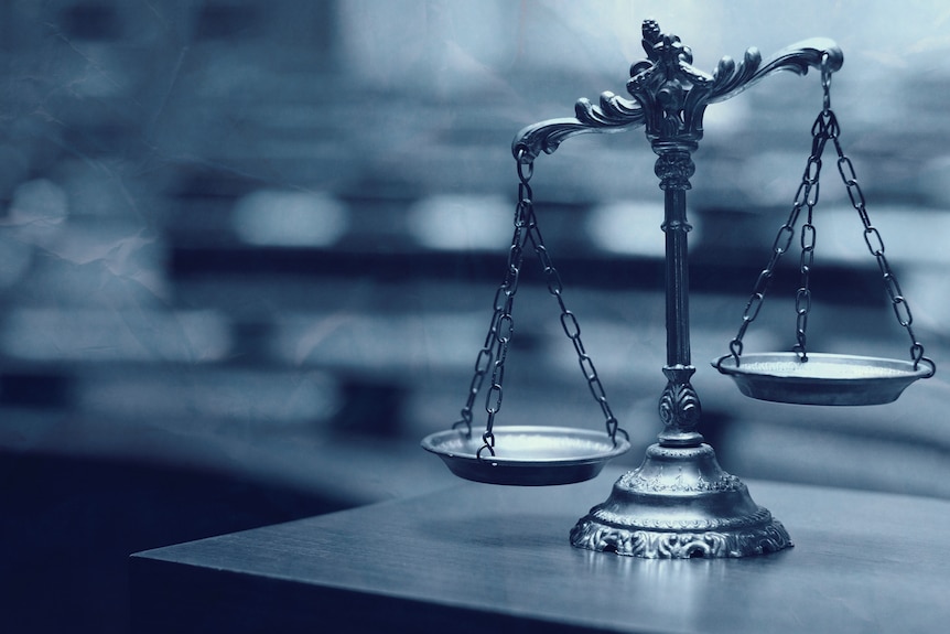 A model scales of justice rests on top of a table, behind it is an out of focus courtroom