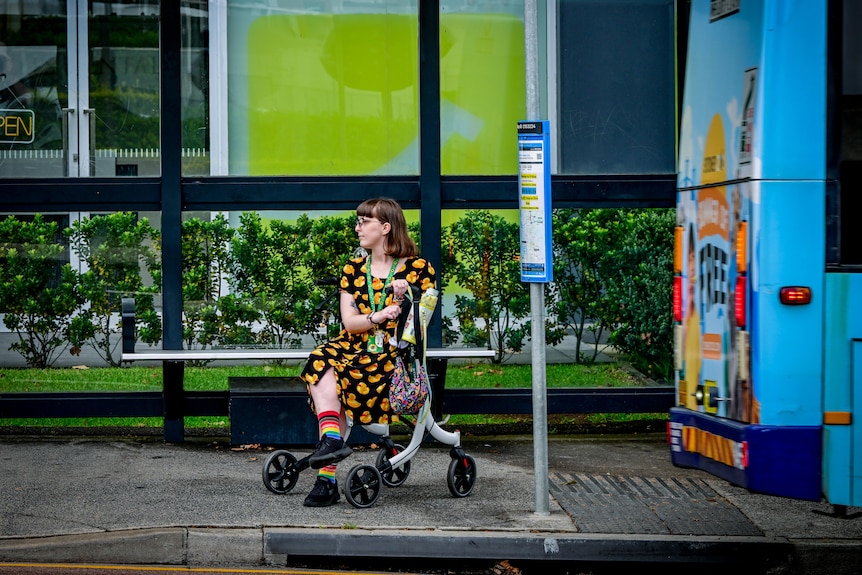 A young woman in a colourful dress sitting on her walking frame. She is on a footpath and has dark brown hair and glasses