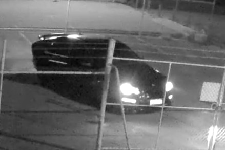 A black and white CCTV image of a dark car parked behind a fence at night with its lights on.