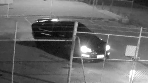 A black and white CCTV image of a dark car parked behind a fence at night with its lights on.