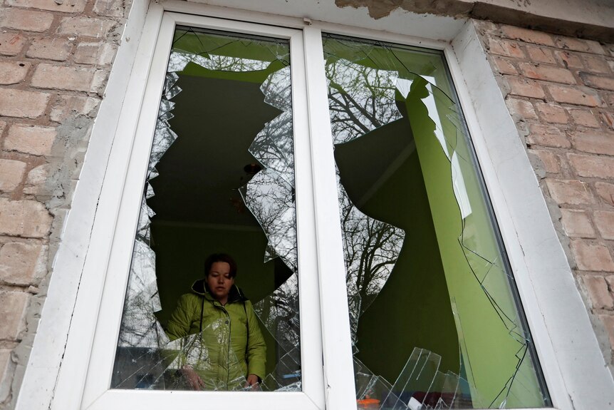 A woman in a yellow jacket stands inside her house, on the other side of shattered windows after Ukraine was invaded by Russia.