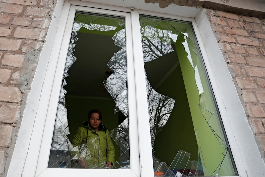A woman in a yellow jacket stands inside her house, on the other side of shattered windows after Ukraine was invaded by Russia.