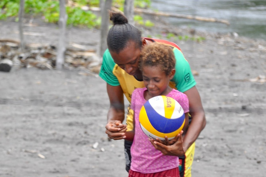 Young girl hold volleyball in left hand as older woman stands behind her, cradling her arms.