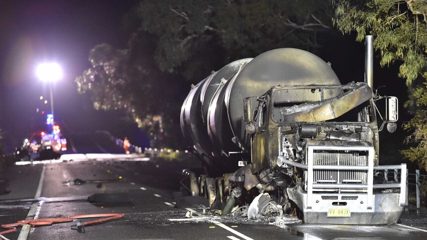A burnt out tanker on the road at night with emergency services in the background.
