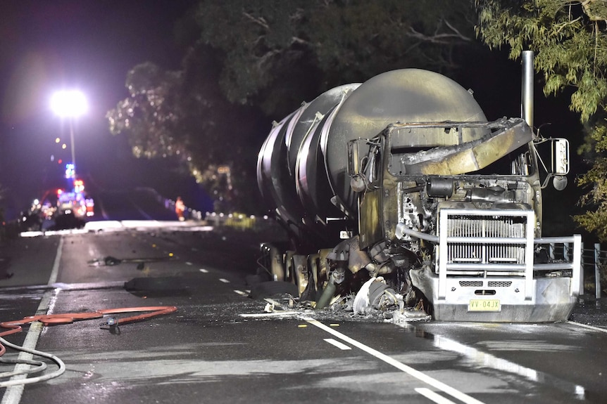 A burnt out tanker on the road at night with emergency services in the background.
