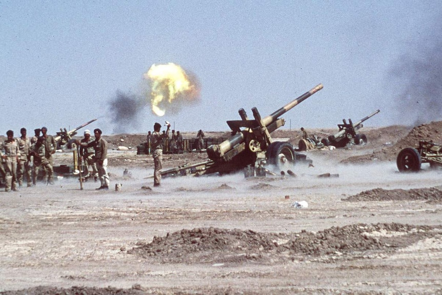 Men in uniform stand around artillery. There is an explosion in the sky above them.