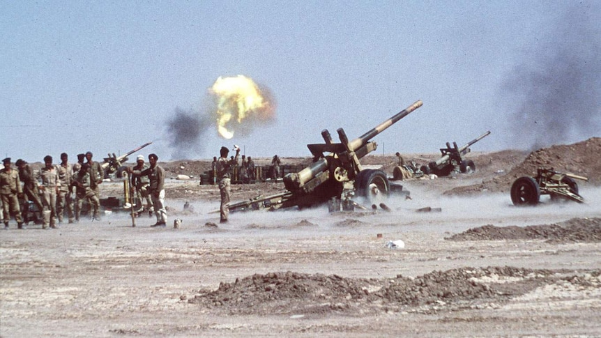 Men in uniform stand around artillery. There is an explosion in the sky above them.