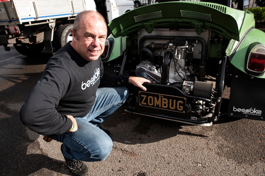 A man crouches by the open hatch of his car with an engine inside