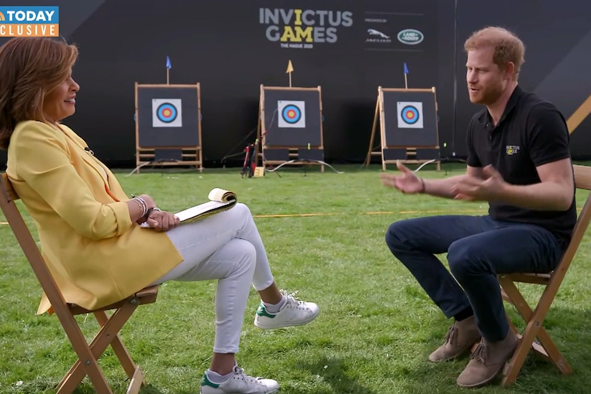 Hoda Ktob and Prince Harry sit on chairs on a grassy area in front of Invictus Games signage. 
