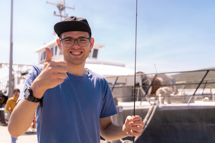 Fishability member Liam stands near a boat, holding a fishing rod in one hand and giving a thumbs up with the other