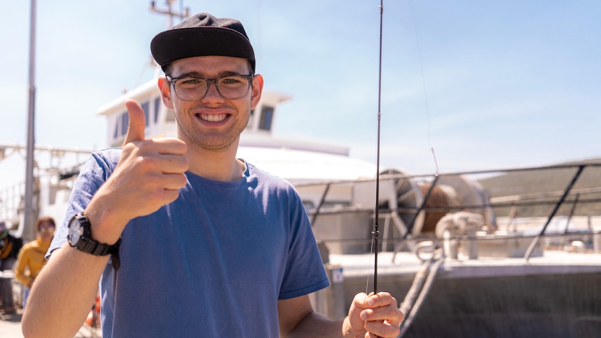 A young man wearing a baseball cap and holding a fishing line gives a 'thumbs up' to the camera.