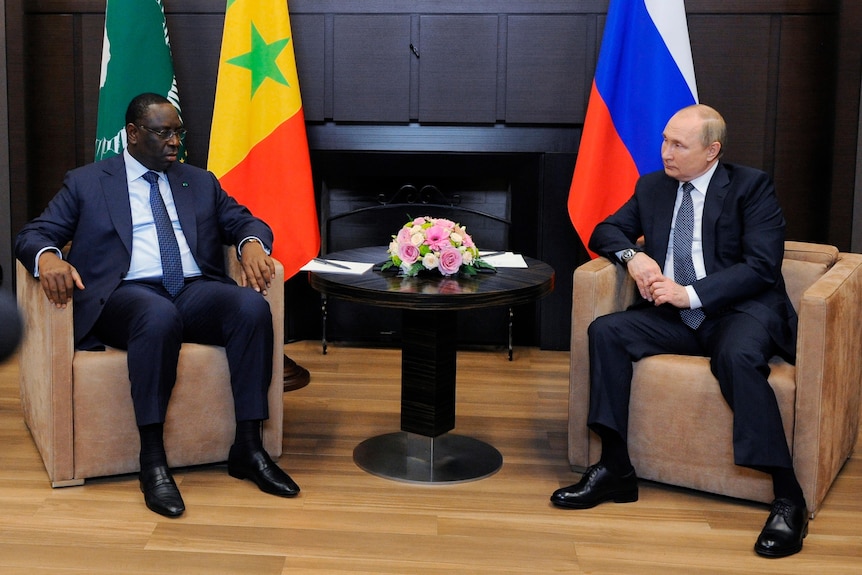 Two men sit at a meeting in front of a Senegalese and Russian flag