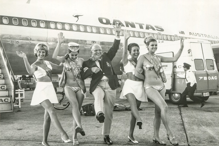 man standing infront on Qantas plane with four women in bikinis