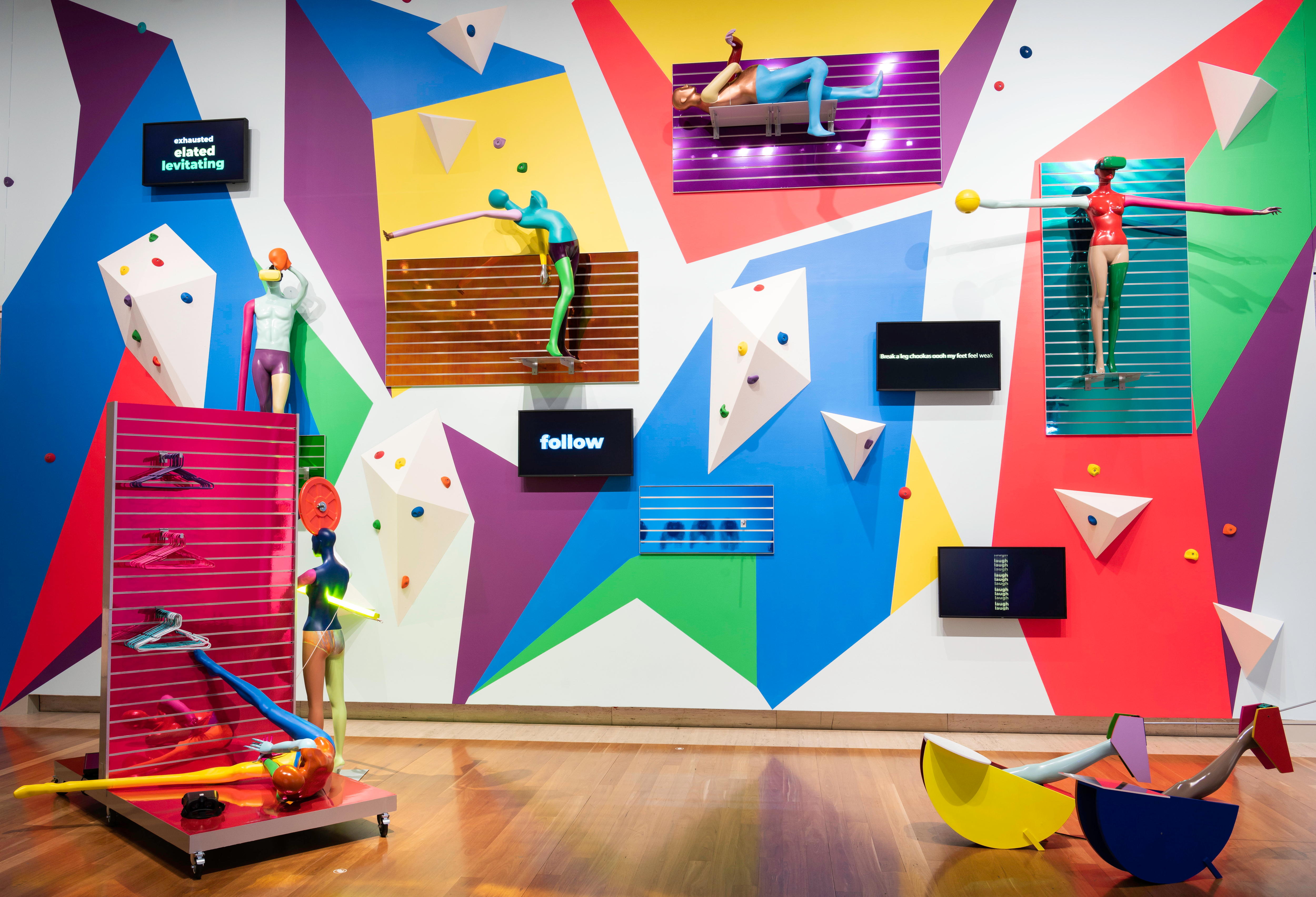 A group of mannequins, painted brightly and manipulated into strange shapes, are attached to yoga mats on a gallery wall.