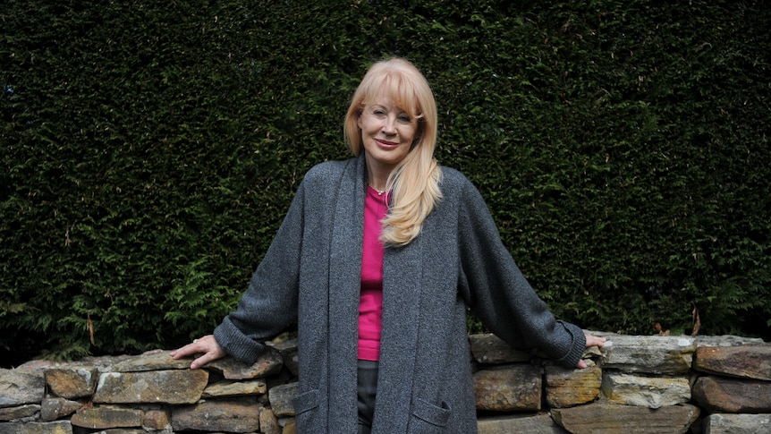 A lady standing in front of a hedge with her hands rested on a low brick wall, smiles towards the camera