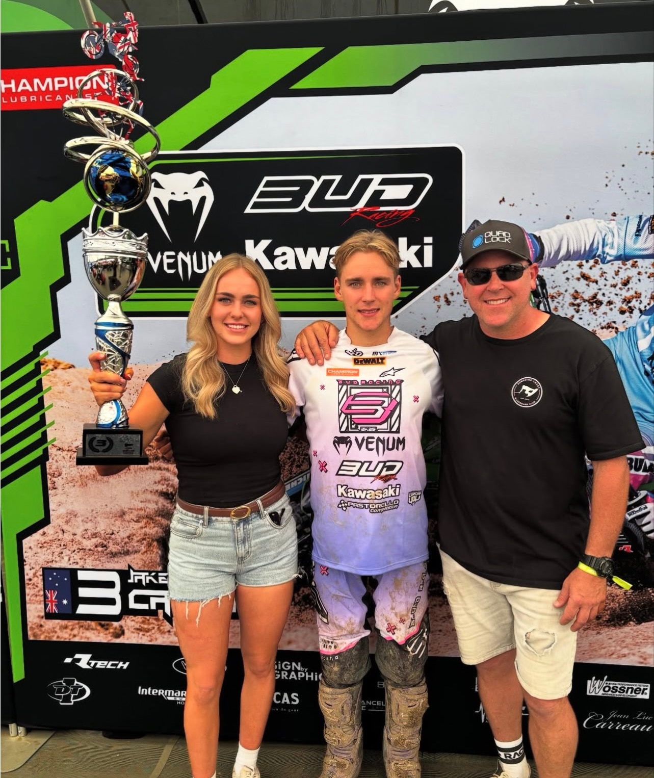 Uma jovem está ao lado de um jovem com equipamento de motocross que também está ao lado do pai.