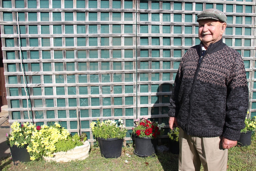 An older man smiles while standing next to potted flowers