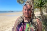 A woman with braids in a colourful shirt smiles at the camera with palm trees and the beach behind her