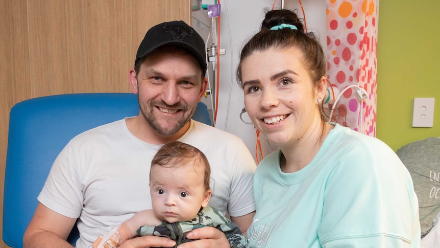 Roger and Rachael smile, holding Leo, who looks surprised and has some tubes attached to his arm.