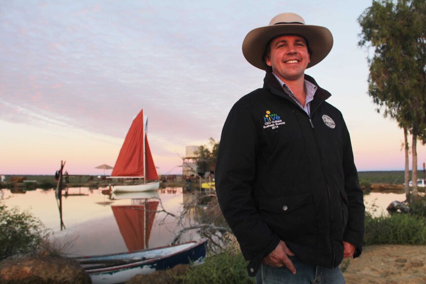 A man in a stockman's hat stands in front of a dam with a boat in the background.