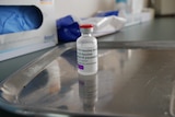 A clear glass vial with a label on a silver tray, with medical gloves and tissues behind