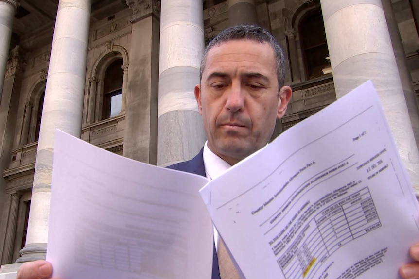Tom Koutsantonis holds pieces of paper in front of his face in front of a building