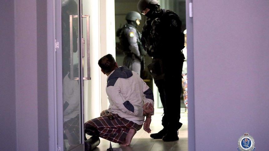 A man crouches on the ground with his hands handcuffed while police look on