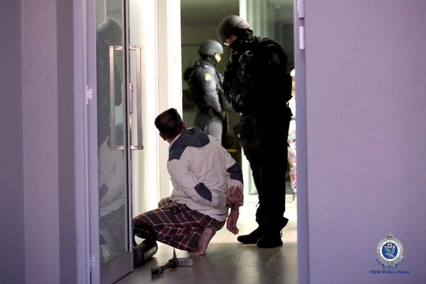 A man crouches on the ground with his hands handcuffed while police look on