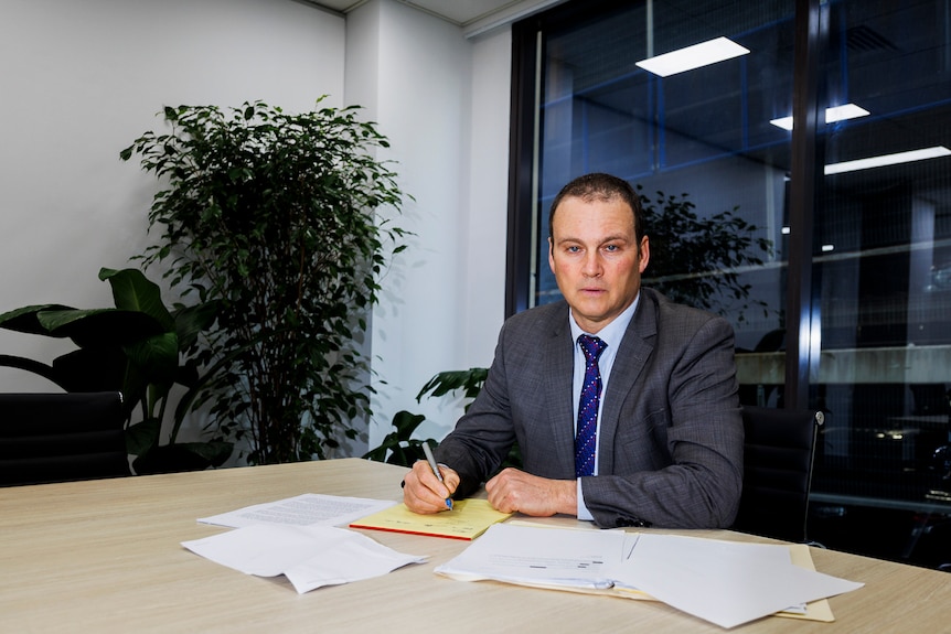 A man in an office sits at a table and turns to face the camera with a serious expression.