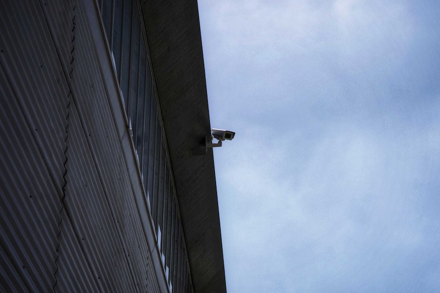 A security camera attached to a high wall points down, behind it is the sky.