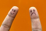 Two index fingers with a happy and sad face drawn on, with orange background.