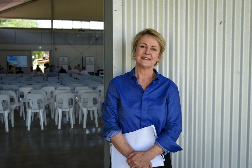 A woman in a blue shirt leans against a silver shed wall with rows of white chairs in the background.