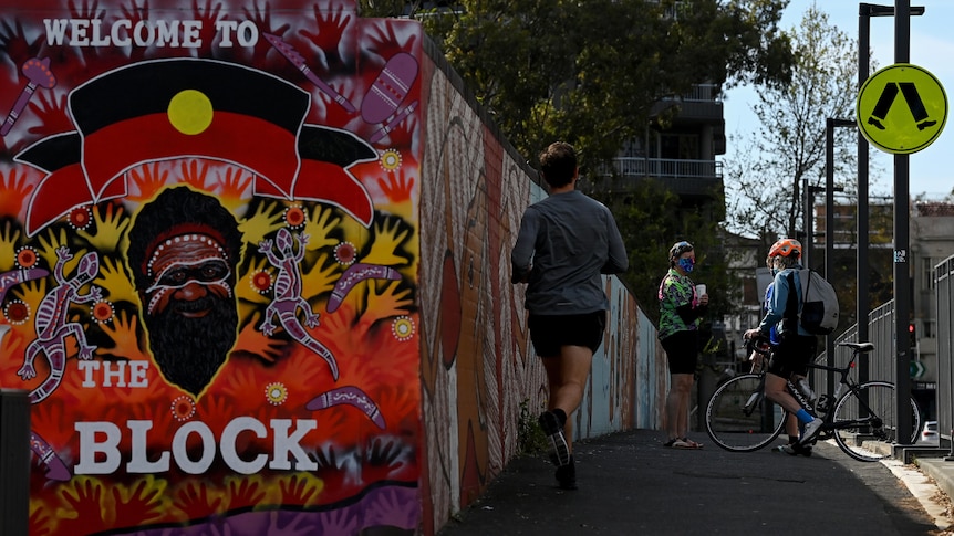 A jogger passing someone standing and talking to a bike-rider near the welcoming sign to Redfern's famous The Block