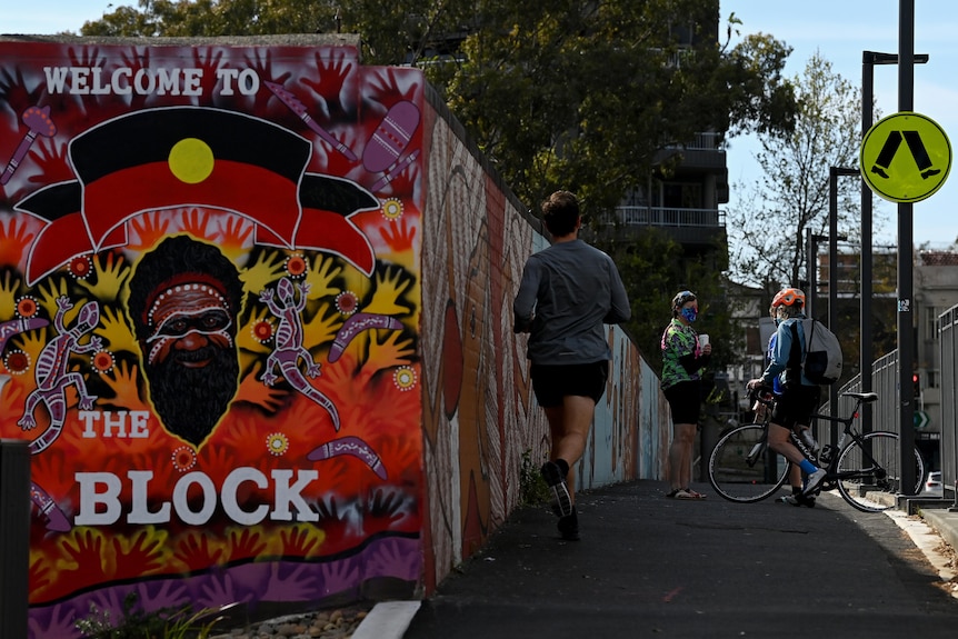 A jogger passing someone standing and talking to a bike-rider near the welcoming sign to Redfern's famous The Block