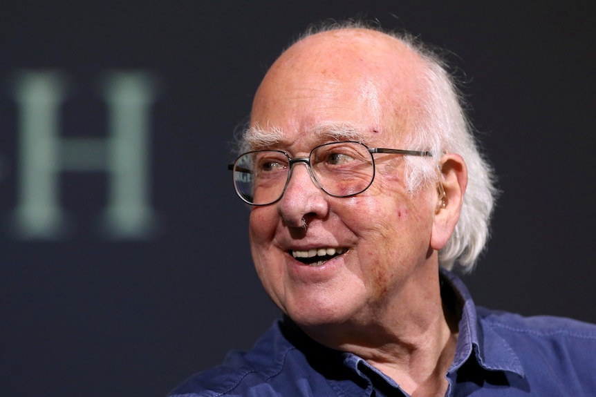 A man wearing round glasses and a blue shirt smiles warmly at someone off-camera