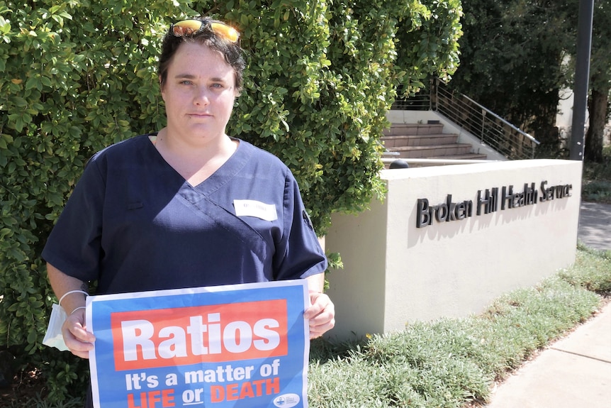 A woman with short, dark, hair, wearing medical scrubs, stands outside holding a protest sign.