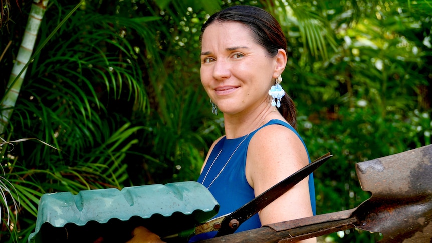 A woman holds garden tools and smiles at the camera. She has raincloud earrings.