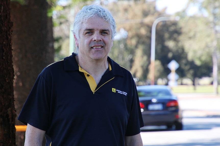 A man wearing a black t-shirt with an L plate on it, with a car in the background.