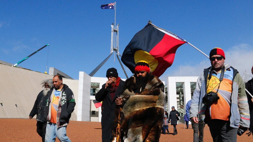 Clinton Pryor, flanked by the Parliament House flag pole and an Indigenous flag.