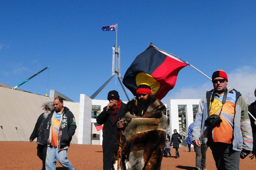 Clinton Pryor, flanked by the Parliament House flag pole and an Indigenous flag.