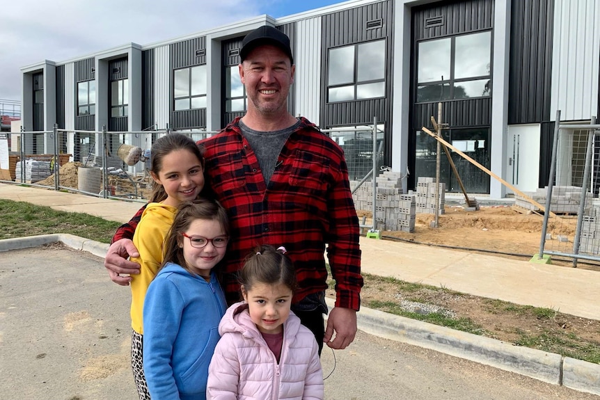 A man stands with his three young daughters in front of a home under construction.