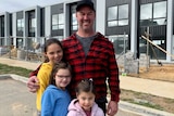 A man stands with his three young daughters in front of a home under construction.