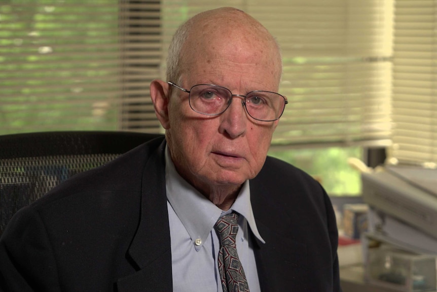 A man in a suit and glasses sitting at his desk with a serious expression