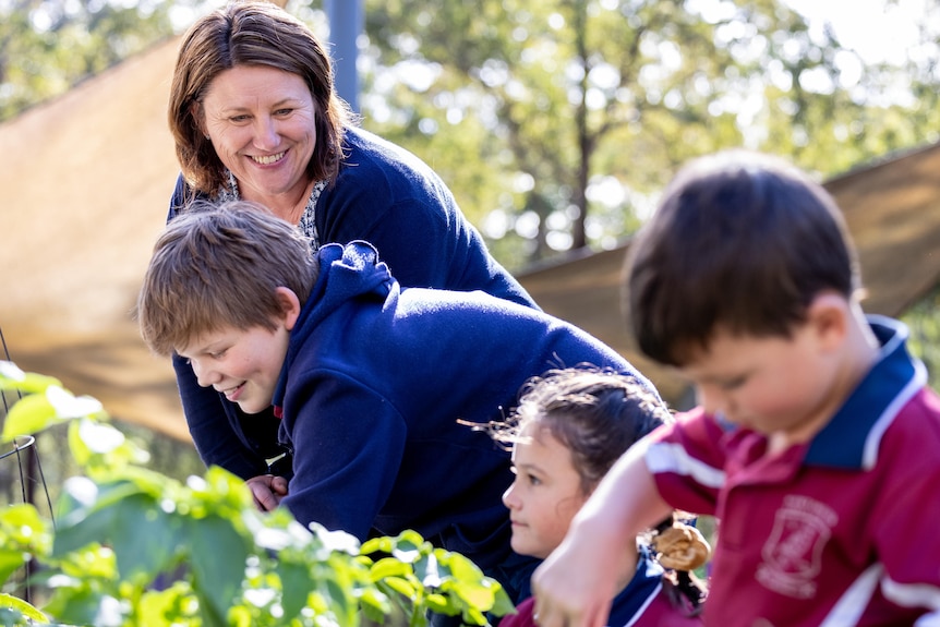 Two boys and a girl tend a garden bed at a primary school, with a teacher standing near them.